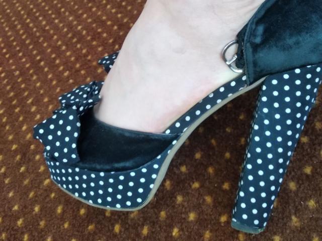 Nurris Fuß in einem schwarzen, gepunkteten High-Heel-Schuh mit einer großen Schleife und einem silbernen Knöchelriemen, der helle Haut und ein leicht sichtbares Sprunggelenk zeigt..