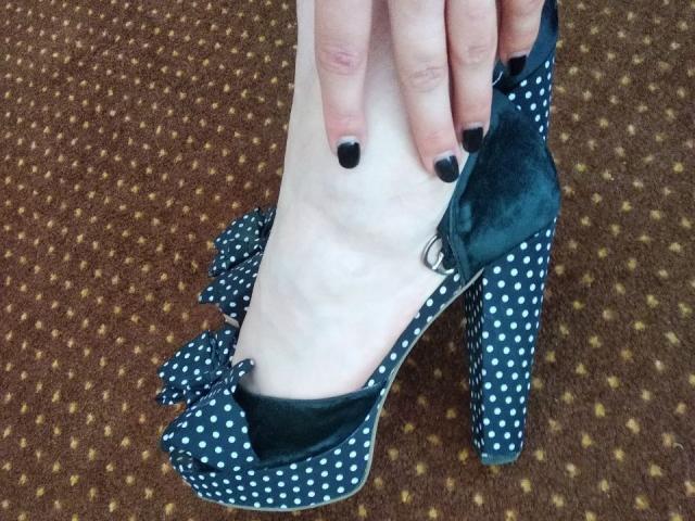 Nurri's pale foot in a blue polka dot high heel with a teal inner lining, showing black nail polish, is being pulled on by Nurri's hand.
