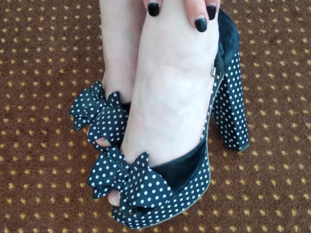 Le pied pâle de Nurri, chaussé d'une haute talon noir à pois avec un grand nœud, son vernis à ongles noir, et le tapis marron avec des points jaunes..