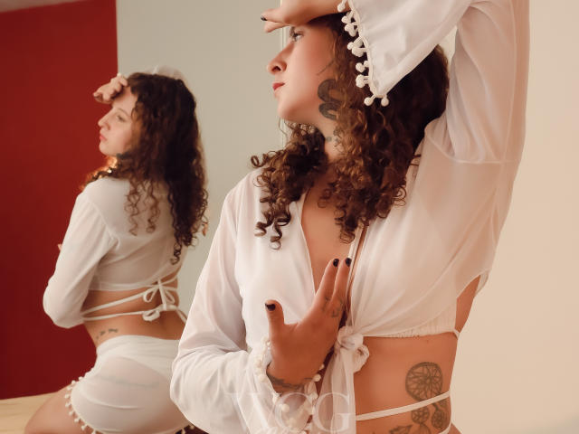 EvaDaMore with curly brown hair, white sheer top, and white strappy bottoms, hand on chest, showing tattoos, looking to the side, reflection in mirror, hands on head.