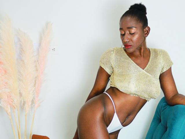 NahomyHawk has dark skin, red lipstick, and a gold crop top, showing her tits and white panties, leaning on a teal chair with puffy pinkish grass.