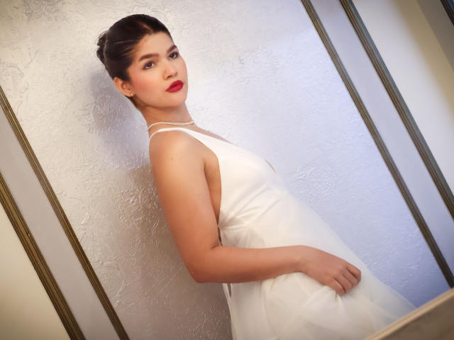 ScarlletZhen mit rotem Lippenstift, weißem Kleid und hochgesteckten Haaren, steht, blickt zurück, zeigt ihre Brüste, hat eine Hand auf ihrem Hintern, vor einer strukturierten Wand, in einem sanft beleuchteten Raum..