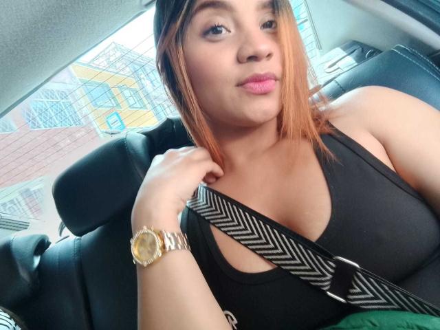 AmbeerBloom with light brown skin, black tank top, small to medium tits, seatbelt, gold watch, pink lips, and orange hair, sitting in a car, slightly pouting.