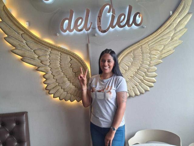 PauletteLoveX with medium tits, smiling, wearing a gray t-shirt, blue jeans, and a tattoo on her right arm, making a peace sign in front of golden wing decor.