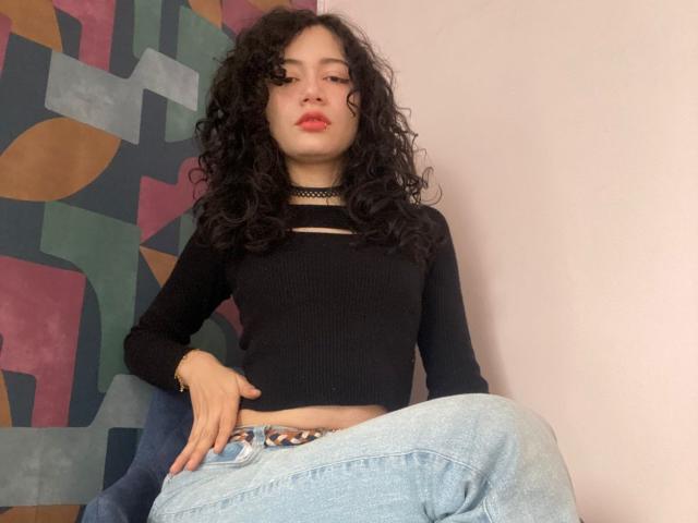 AmeliaTorres with curly black hair, red lips, black choker, black long-sleeve top, and light blue jeans, sitting with hand on hip, looking slightly annoyed.