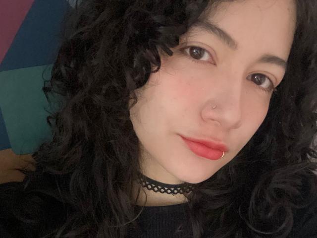 AmeliaTorres has curly black hair, a black choker, red lipstick, and nose piercings. She's looking at the camera with a slight smile. Her tits are not visible.
