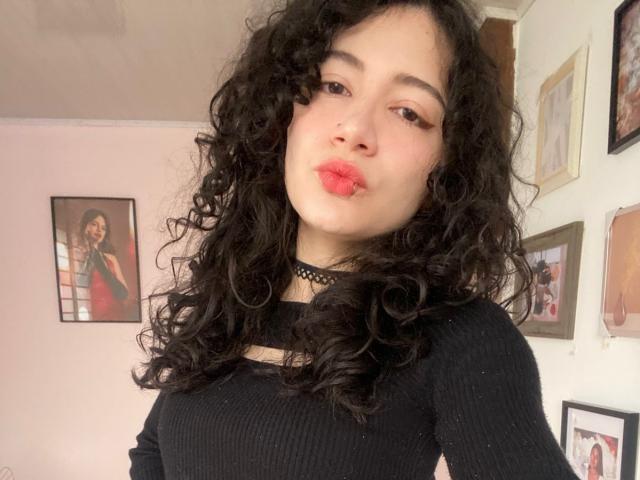 AmeliaTorres with curly black hair, pouty red lips, wearing a black choker and ribbed black top, looking at the camera. Boobs not visible, background photos in the room.