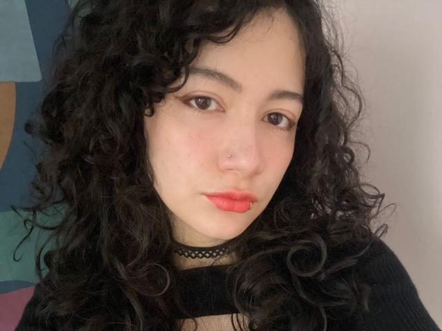 AmeliaTorres has curly black hair, a black choker, red lipstick, and a nose ring. She's looking at the camera with a slight, confident smile. Her tits are small.