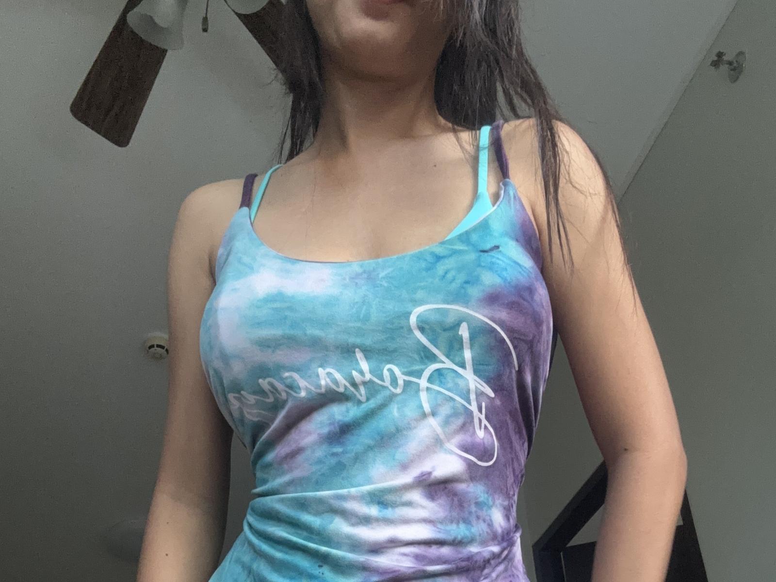 Ressy with her tits in a blue and purple tie-dye tank top, showing some cleavage, standing indoors, arms at sides, long dark hair, partially visible ceiling fan above.