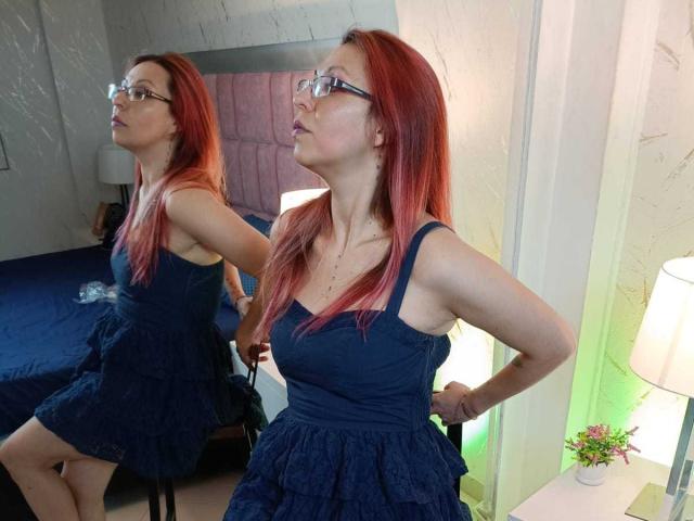ArtemisFlyve with red hair, glasses, and a navy blue dress, pouting and hands on hips, reflected in a mirror. Boobs visible in dress, small to medium size.