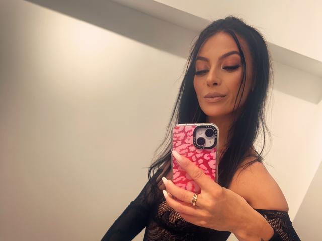 KinkyAmelie with long black hair, wearing a black lace off-shoulder top, holding a pink phone with white patterns, taking a selfie, showing her subtle makeup and gold ring.