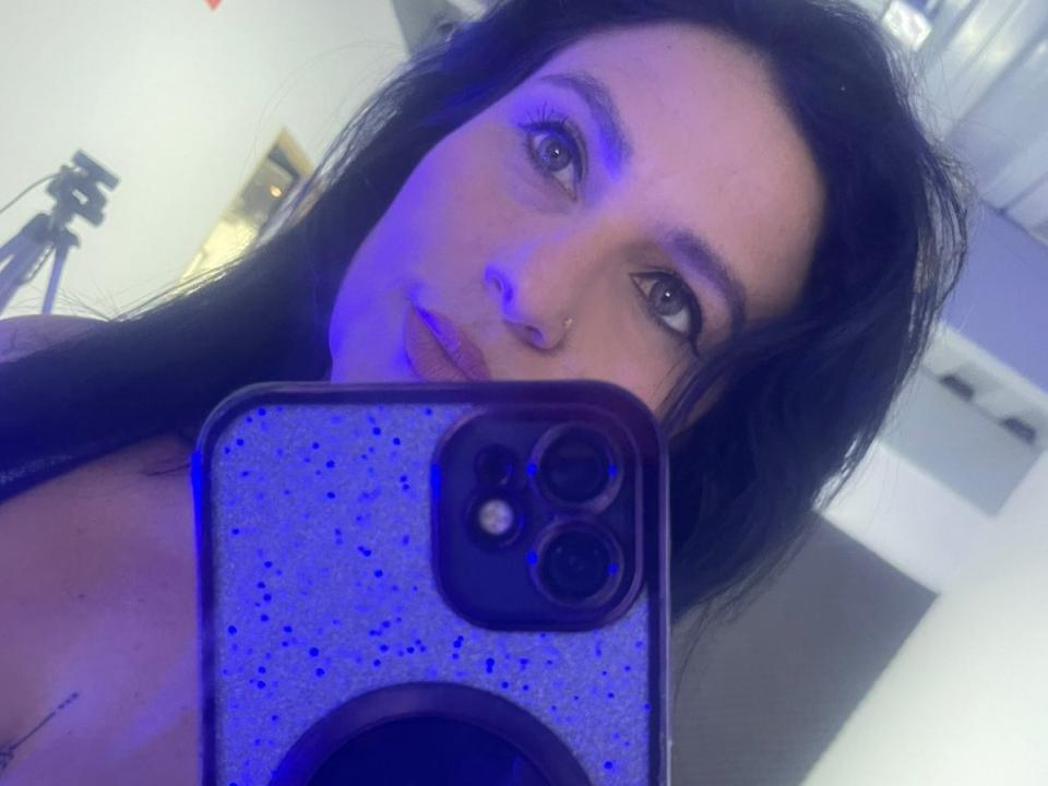 NadiaSamiri with long black hair, blue-tinged face, nose ring, holding a purple speckled phone, covering her tits, taking a selfie with a camera tripod in the background.