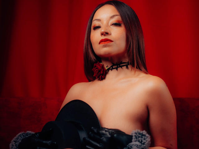 AngelinaBlanche with bare boobs, wearing black gloves and a black choker with a rose, has red lipstick and a confident, sexy expression against a red curtain.