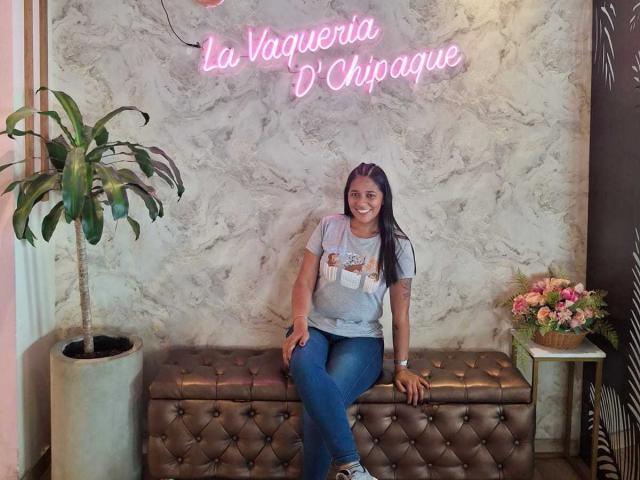 PauletteLoveX with long dark hair, smiling, wearing a gray tee and blue jeans, sitting on a brown tufted bench, potted plant on left, pink neon sign above.