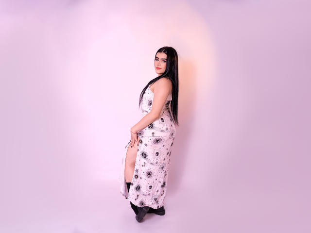 VynxDarkFire stands in a pink-lit room wearing a white floral dress with a thigh slit, black socks, and black heels, looking over her shoulder with long black hair.