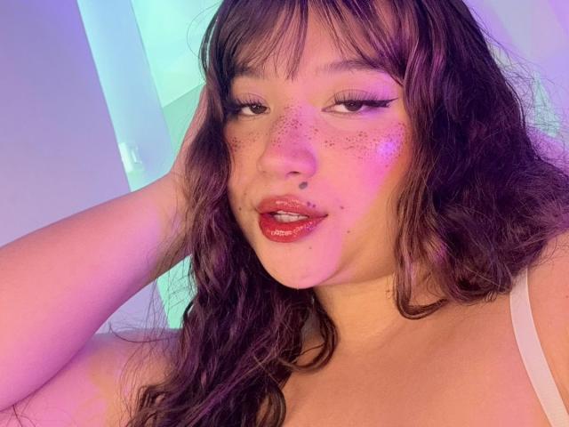 HonnyBlaice with wavy brown hair, freckles, and red lipstick, wearing a white top, has her hand in her hair, looking seductively at the camera with pink and purple light.