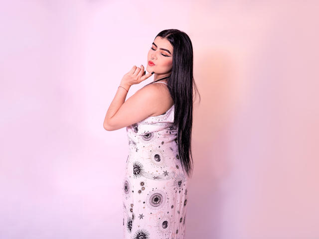 VynxDarkFire with long black hair, wearing a white dress with black star patterns, standing sideways, eyes closed, touching her chin, pink and purple background.