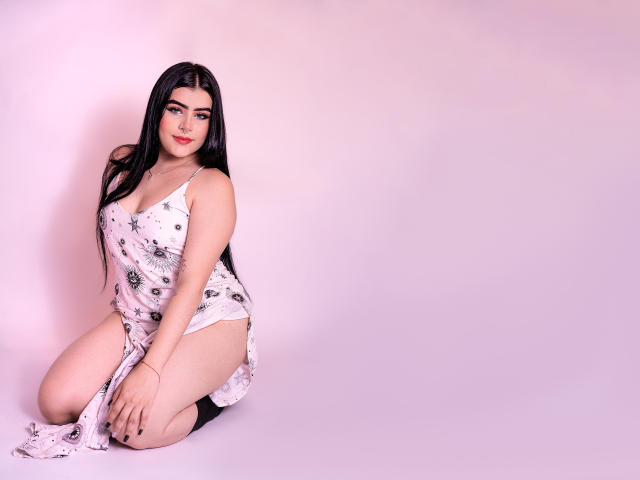 VynxDarkFire, kneeling, wears a white floral dress, showing her ass, with long black hair, red lipstick, and black stockings, against a pink background.
