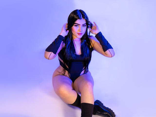 VynxDarkFire with long black hair, wearing a black one-piece, black wristbands, and black socks, sitting with hands in hair, blue and purple lighting. Boobs visible, ass prominent.
