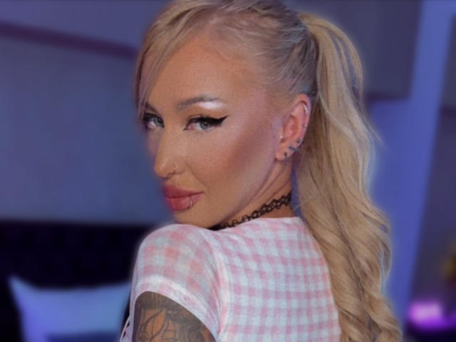 SinnersMuse has blonde hair in a ponytail, pink checkered shirt, tattooed arm, black choker, and nose ring, with heavy eyeliner and a pouty expression.