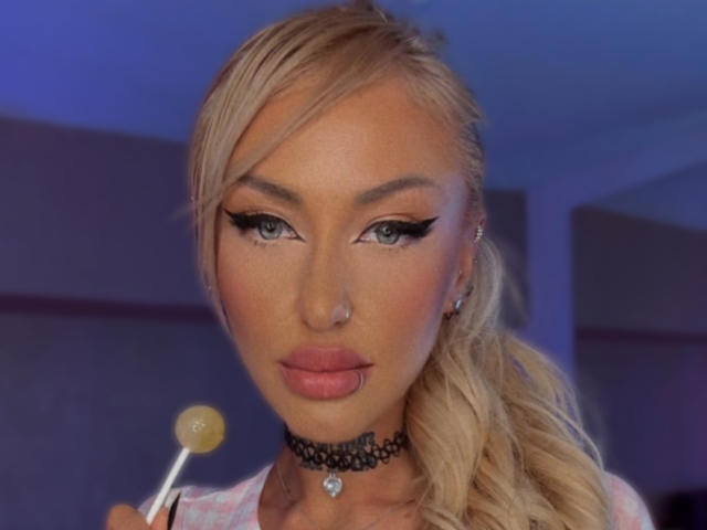 SinnersMuse with blonde hair, blue eyes, and black eyeliner, holding a yellow lollipop, wearing a black choker, and pink plaid shirt. Her ass is not visible.