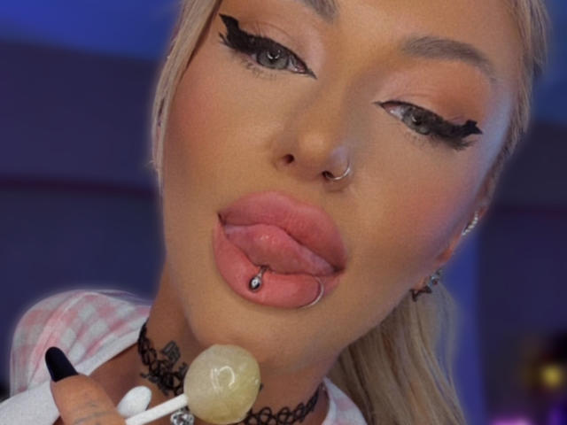 SinnersMuse with blonde hair, black eyeliner, nose ring, and pink tongue piercing, sucking a lollipop, has a lip piercing and choker, and is looking up with her mouth open.
