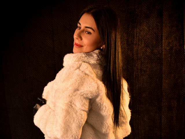 Maddy Smit, met lang bruin haar, een witte, pluizige jas en rode lippenstift, lacht naar de camera, houdt zwarte handschoenen vast, donkere achtergrond, warm licht, zachte textuur, aantrekkelijk, zelfverzekerd..