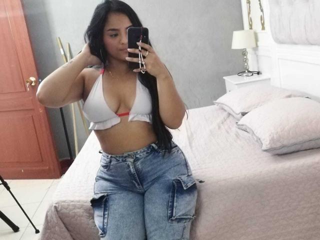 RileyPrescott with long black hair, white bikini top showing small tits, pink nipples, and high-waisted blue jeans, taking a mirror selfie in a bedroom.