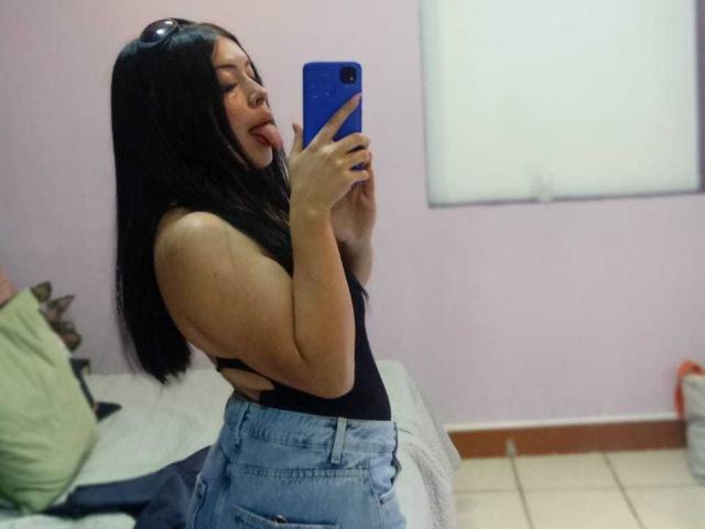 KateWelsin with long black hair, tongue out, wearing a black top and blue jeans, taking a selfie in a mirror, holding a blue phone, in a pink-walled room.