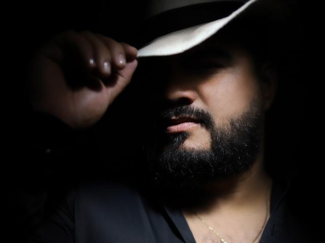 El rostro de Michael Baer, con barba oscura y parcialmente en sombra, sostiene el borde de un sombrero blanco, con los ojos cerrados, vistiendo una camisa negra y con un discreto collar dorado, en una fotografía con poca luz..