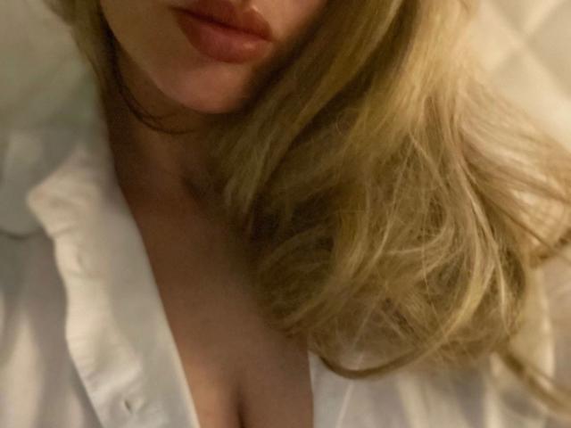 Kathinka-hot's face with blonde hair, slightly parted lips, and a white shirt, showing a bit of cleavage. Her tits are partially visible. The image is a photo.