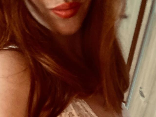 Kathinka-hot's red lips and long, wavy red hair, with a peek of her lace-covered boobs, in a slightly blurred, intimate selfie.