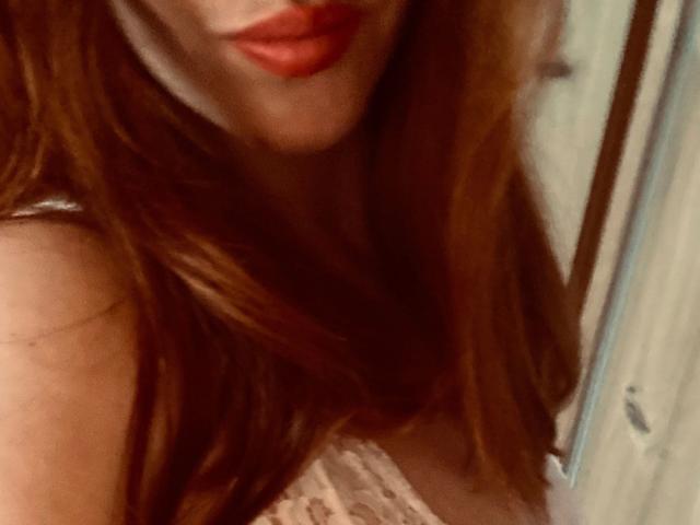 Kathinka-hot with red lipstick, partial view of her face and chest, showing her tits in a lace top, long red hair, slightly blurry photo.
