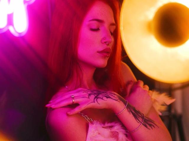 CherrySmarry-hot with red hair, nose ring, and tattooed hand, in pink and yellow neon light, wearing fluffy white top, hands near chest, eyes closed, subtle smile.
