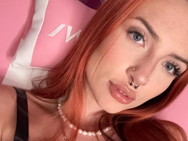 CherrySmarry-hot has red hair, blue eyes, and multiple nose piercings with black dots. She's wearing a pearl necklace and has glossy pink lips. Her black strap is visible.