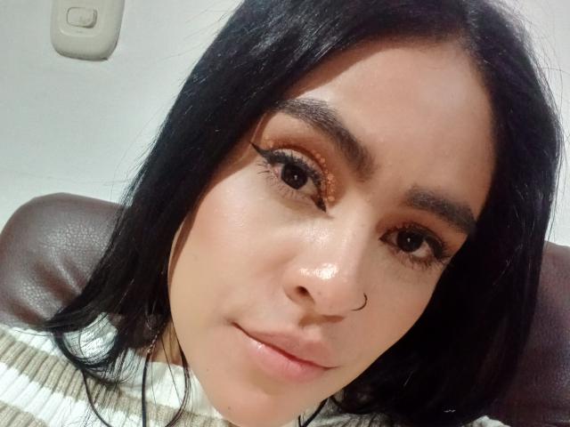 StellaBeckker has tan skin, black hair, and brown eyes with bold eyeliner. She's wearing a nose ring and a striped shirt, with a slight smile on her face.