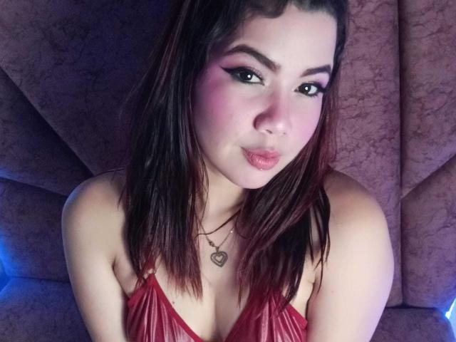 Hillary Riley, con rubor rosado, vestido rojo, collar con forma de corazón, labios fruncidos y cabello castaño oscuro, mirando a la cámara, mostrando sus senos. Sus ojos están delineados con delineador negro..