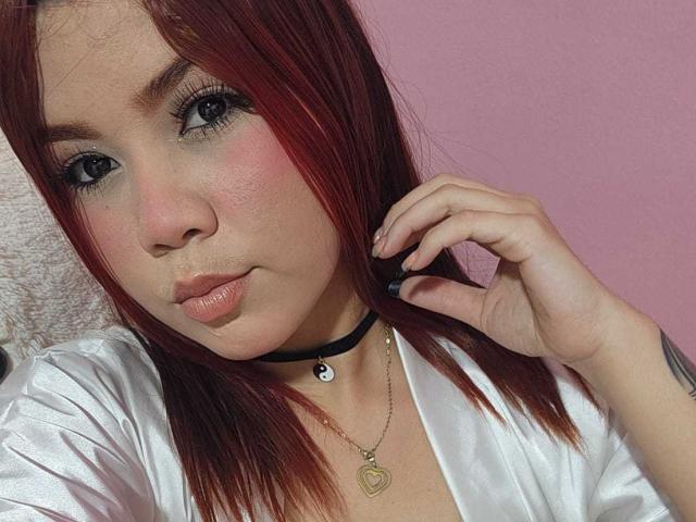 Hillary Riley hat rotes Haar, blaue Augen und rosigen Lippen. Sie trägt eine schwarze Halskette, eine weiße Satinbluse und eine goldene Herz-Kette, und ihre Hand ist in der Nähe ihres Gesichts..