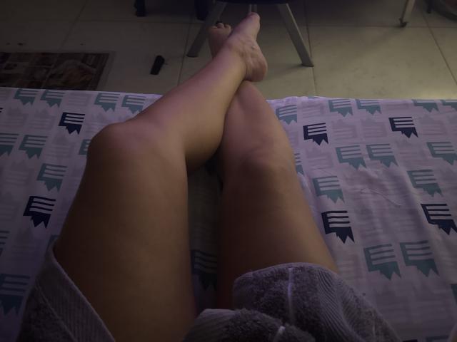 SeleneBrigth's legs crossed on a bed, showing her bare thighs and feet, wearing a white towel, dimly lit, with a chair and tile floor in the background.