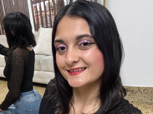 DulceSweety, with black hair and pink eyeshadow, smiling at the camera. She's wearing a black lace top, and her ass is visible in the mirror reflection.