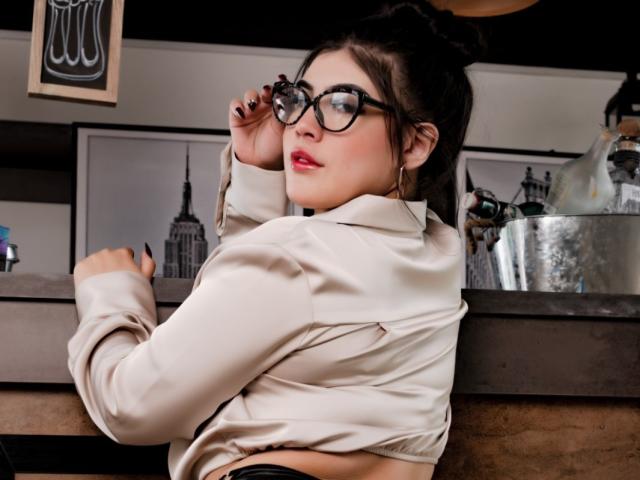 SweetEmily69 with glasses, red lips, and black hair in a bun, wearing a beige shirt, leaning on a counter, looking over her shoulder, showing her tits.