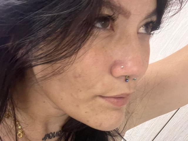 VanellopeWow has a nose piercing with blue and green jewels, long dark hair, and a slight scar on her right cheek. Her brown eyes are partially closed.