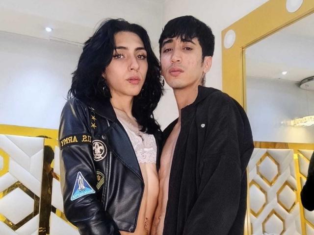 AsmodeusLilith with black wavy hair, wearing a black leather jacket, showing bare tits, stands next to AsmodeusLilith with short black hair, black shirt, and exposed chest.