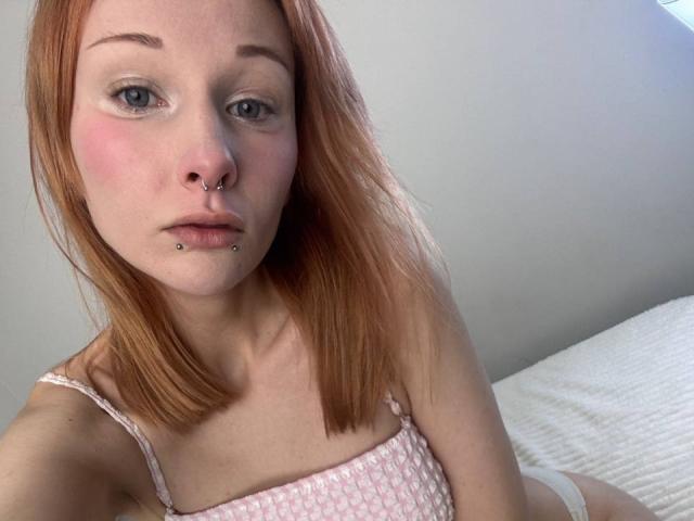 HannaCool has red hair, blue eyes, and a nose ring with a lip piercing. She's wearing a pink, patterned top, and white underwear, with a white bed behind her.