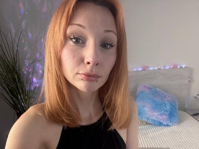 HannaCool with red hair, nose ring, lip piercings, black top, and eyeliner, sitting in a bedroom with a blue fuzzy pillow and string lights in the background.