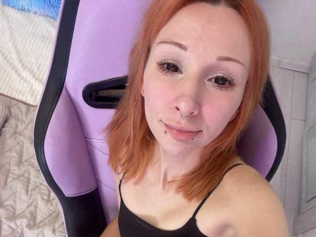 HannaCool, con cabello rojo, aro en la nariz y piercing en el labio, vestida con una camiseta negra, sentada en una silla de juego morada y negra, sonriendo a la cámara..