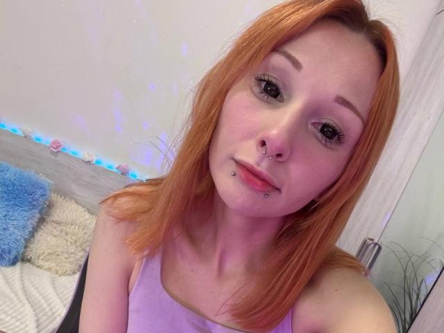 HannaCool has red hair, nose and chin piercings, wearing a purple top, with a cute smile and blue eyes. Her ass is not visible. She's on a bed.