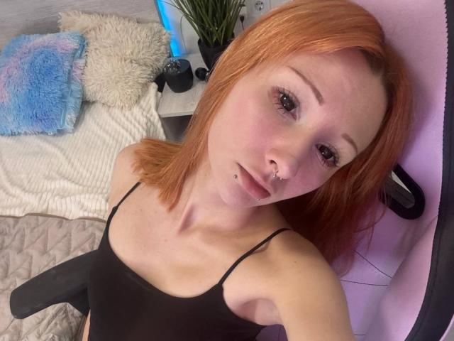 HannaCool with red hair, nose piercing, and black top, sitting in a chair, showing small tits, slight pout, and a lip piercing, looking at the camera.