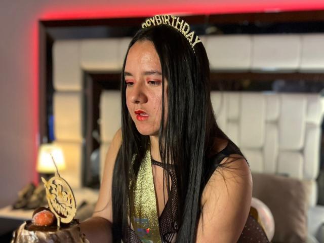 SaraFountain with long black hair, wearing a "Happy Birthday" tiara, fishnet top, red lipstick, holding a chocolate cake with a candle, in a dimly lit room.