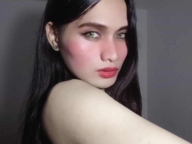 AmberH with pale skin, green eyes, pink blush, red lips, black hair, and earrings, looking over her shoulder, showing her bare shoulder and upper back.